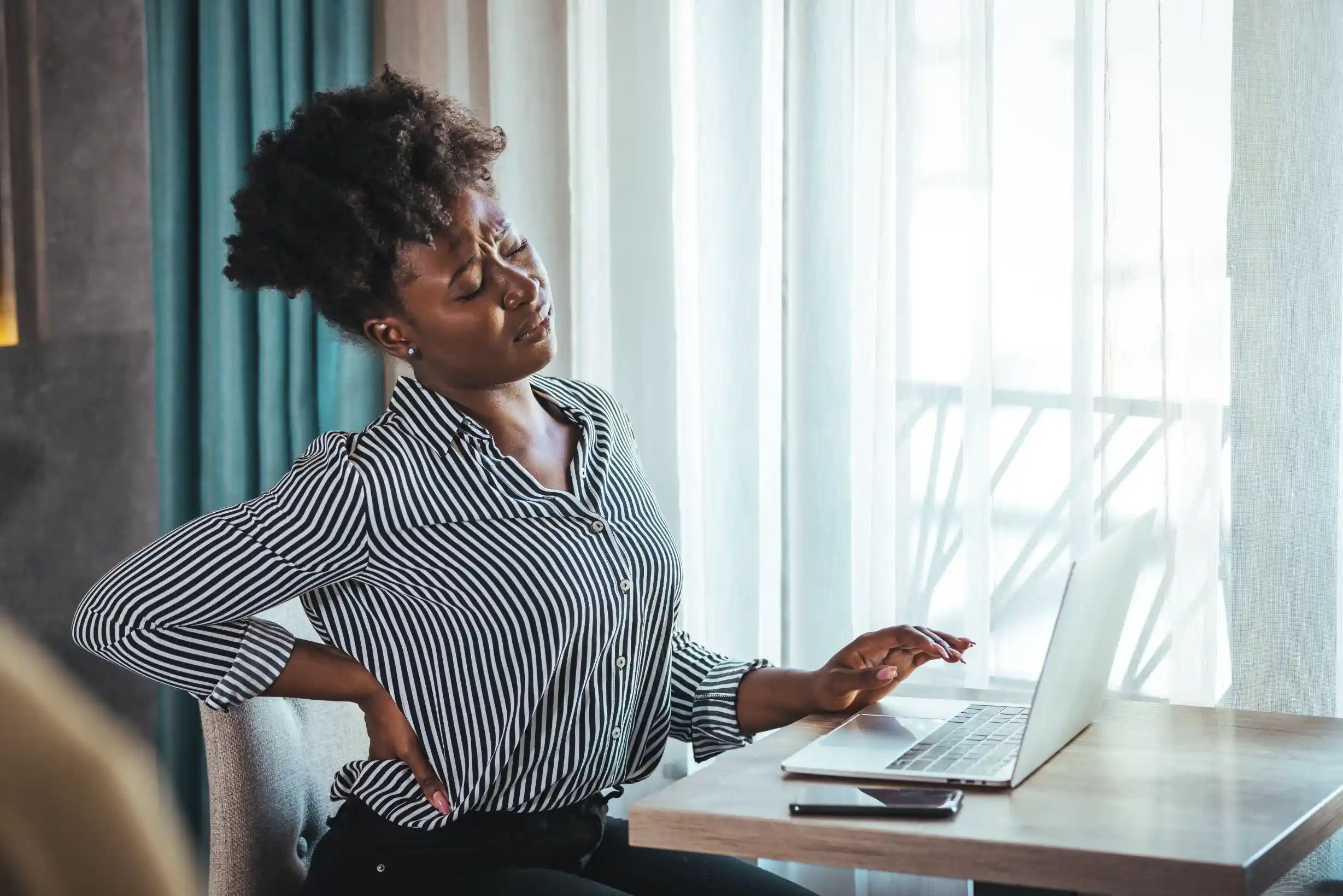 A woman holding her back painfull while working on her laptop.