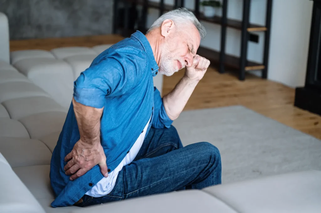 Man sitting on couch holding lower back with a pained expression.