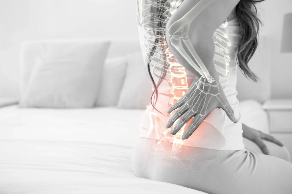 Woman sitting on a bed with hands on lower back, overlaid with a glowing illustration of her spine to depict spinal cord pain and the potential need for spinal cord stimulation.
